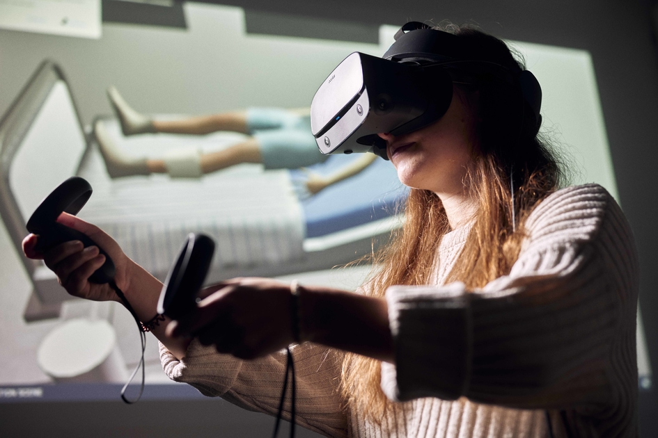 Eine Frau mit VR-Brille und je einem Controller in jeder Hand. Im Hintergrund eine Leinwand, die einen auf dem Bett liegenden Patienten zeigt.