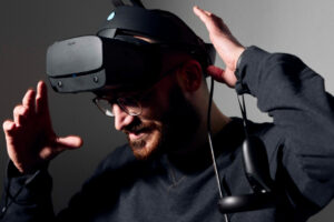 Ein dunkel gekleideter Mann mit Bart und Brille, dem die Controller schon an den Handgelenken hängen, führt seine Hände zum Kopf, um sich die VR-Brille über die Augen zu ziehen.
