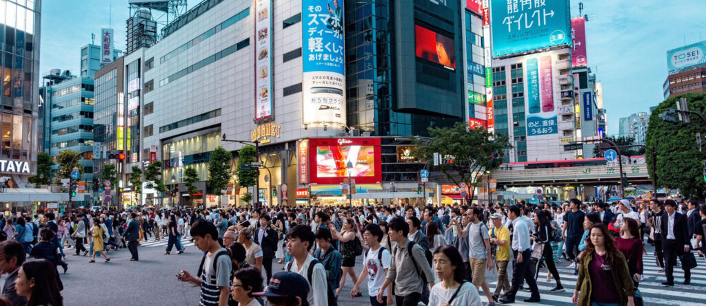 Menschen, die in Tokio über einen Zebrastreifen gehen. Im Hintergrund mehrere Gebäude mit japanischen Werbedisplays.