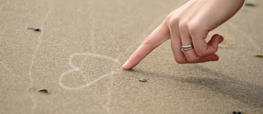 Eine Frauenhand malt mit dem Zeigefinger ein Herz in den feuchten Strandsand.