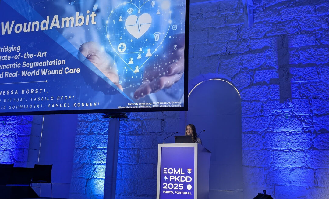 Für ihr Projekt WoundAmbit wurde die Informatikerin Vanessa aus Würzburg auf der der ECML PKDD mit dem “Best Student Paper Applied Data Sciences Track” ausgezeichnet. Blick auf die in blauem Licht erstrahlte Bühne mit Rednerpult und großer Leinwand.