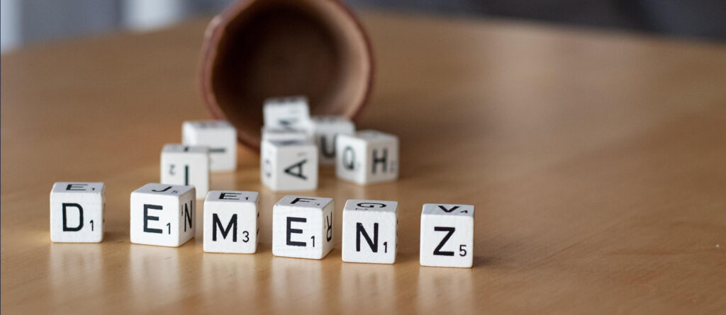 Scrabble-Steine, die aus einem im Hintergrund liegenden Würfelbecher gefallen sind, formulieren das Wort "Demenz".