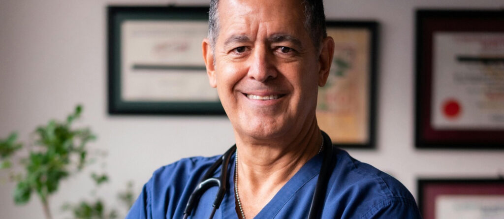 Ein Arzt "in den besten Jahren" lächelt in die Kamera. Im Hintergrund hängen Zertifikate an der Wand.