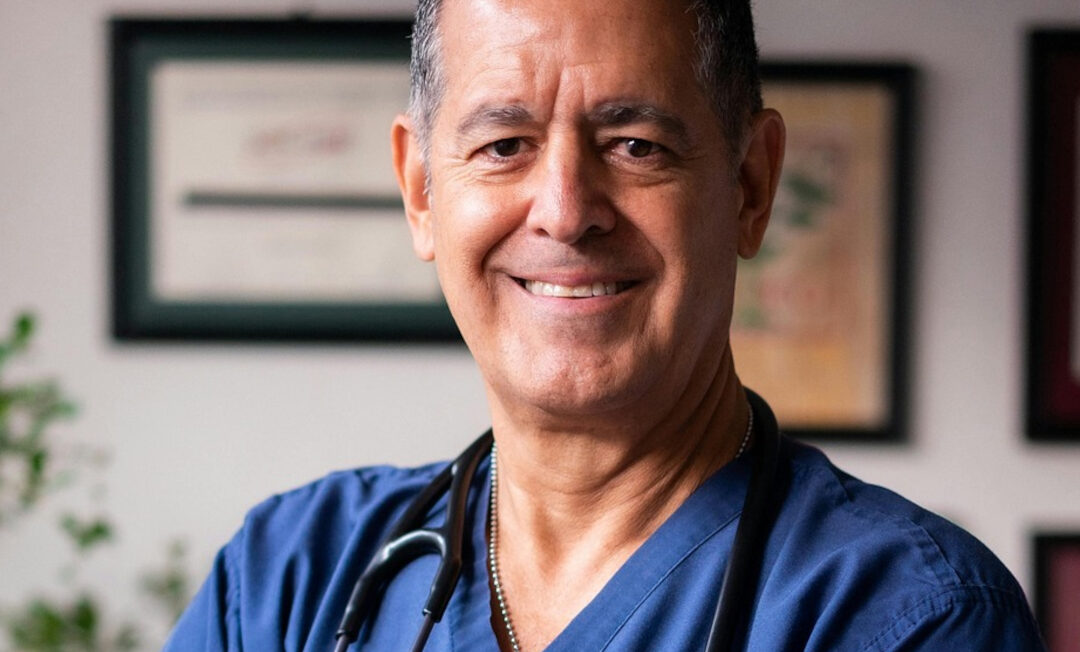 Ein Arzt "in den besten Jahren" lächelt in die Kamera. Im Hintergrund hängen Zertifikate an der Wand.
