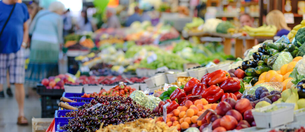 Ein Stand mit Obst und Gemüse. Seitlich im Hintergrund ein paar Kunden.