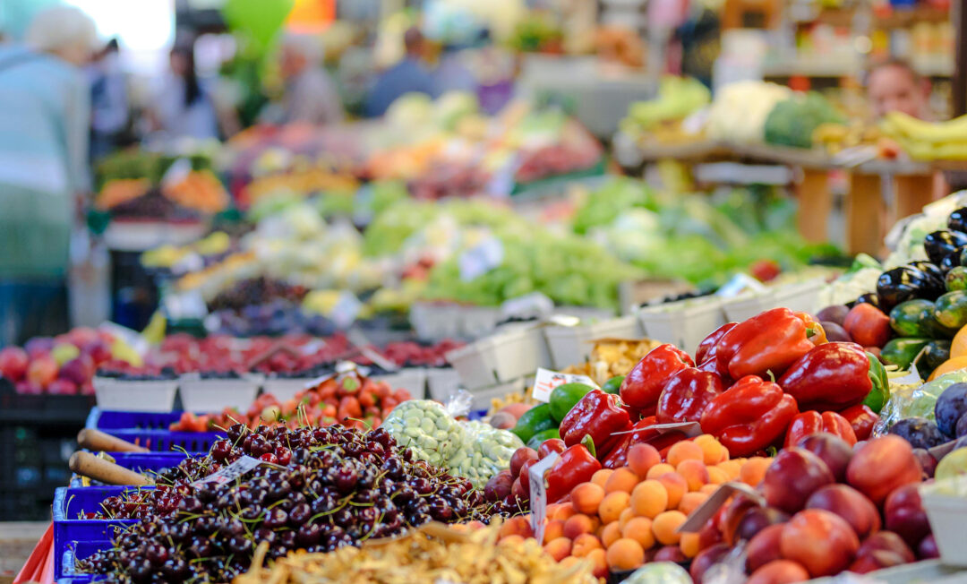 Ein Stand mit Obst und Gemüse. Seitlich im Hintergrund ein paar Kunden.