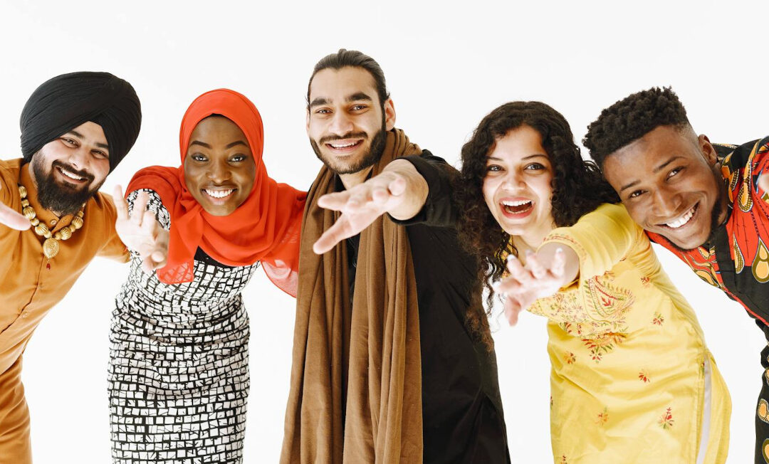 Fünf Personen aus unterschiedlichen Kulturkreisen, teils landestypisch gekleidet, beugen sich, Hände vorgestreckt nach vorne und lächeln in die Kamera. Sie sehen aus, als hätten sie viel Spaß.