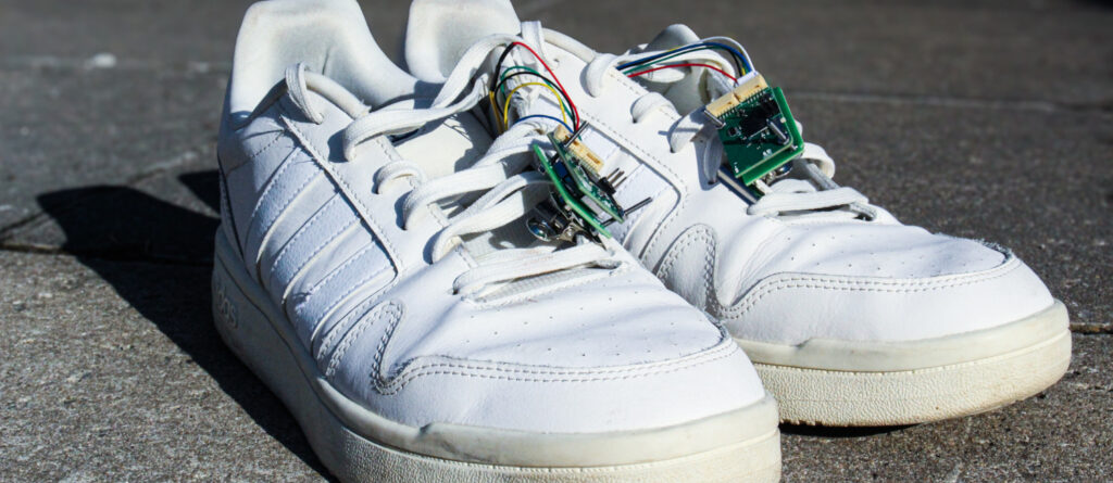 Ein paar Sportschuhe. Am Schnürsenkel hängt eine Platine. auch dünne Kabel sind sichtbar.