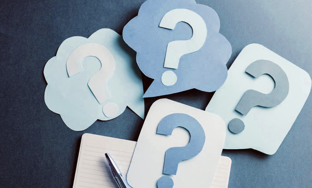 Sprechblasen aus Papierblättern, auf denen Fragezeichen aus Papier liegen.