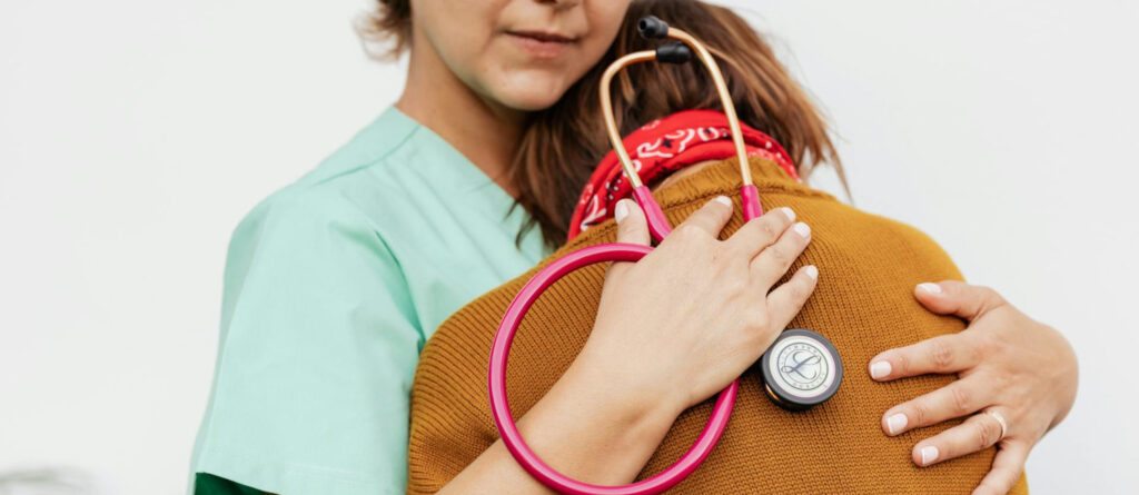 Eine Ärztin oder Pflegekraft mit Stethoskop in der Hand nimmt eine ältere Patientin tröstend in den Arm.